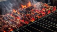 Nikmatnya Sate Klatak Bakar di Atas Bara Arang Sate Klatak sedang dibakar di atas bara api arang yang menyala merah dengan asap mengepul, menunjukkan potongan daging yang ditusuk pada jeruji besi.